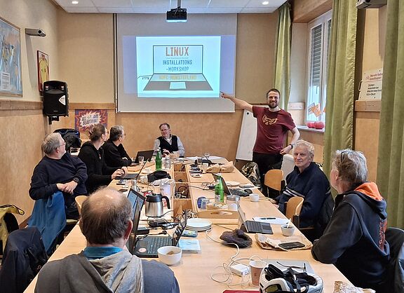Auf dem großen Gruppentisch im Clubraum des ADFC Münsterland stehen viele geöffnete Laptops. An dem Tisch sitzen Menschen. Auf der Leinwand ist eine Zeichnung von einem Laptop zusehen auf dem Bildschirm steht : Linux, Linux-Stammtisch Münster, Installations-Workshop, ADFC-Münsterland. Auf einem USB-Stick steht Linux.