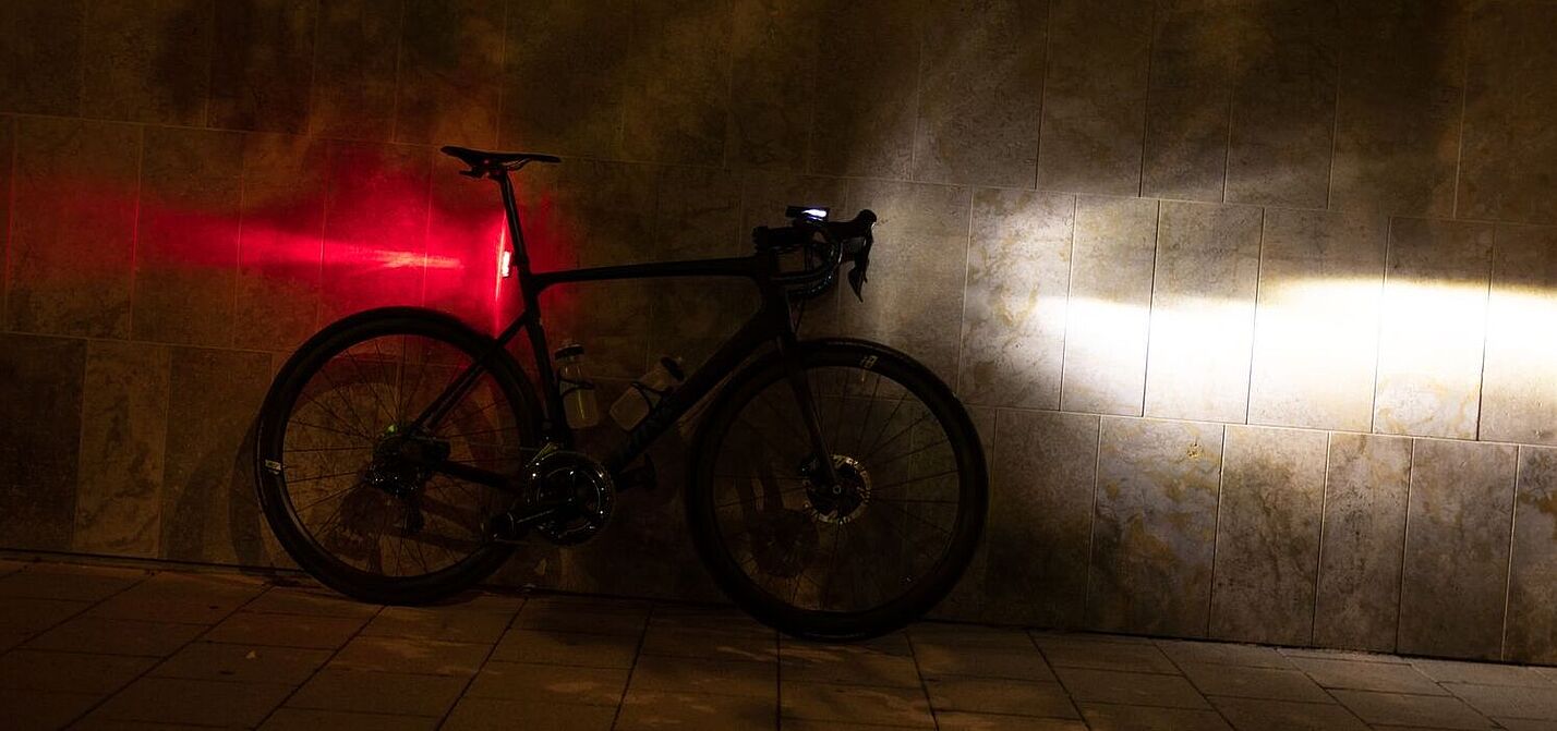 Sigma Aufmacher Ein Fahrrad steht in der Dunkelheit an einer Wand, mit am Lenker montiertem, eingeschalteten Scheinwerfer und an der Sattelstütze montierten, leuchtenden Rücklicht.