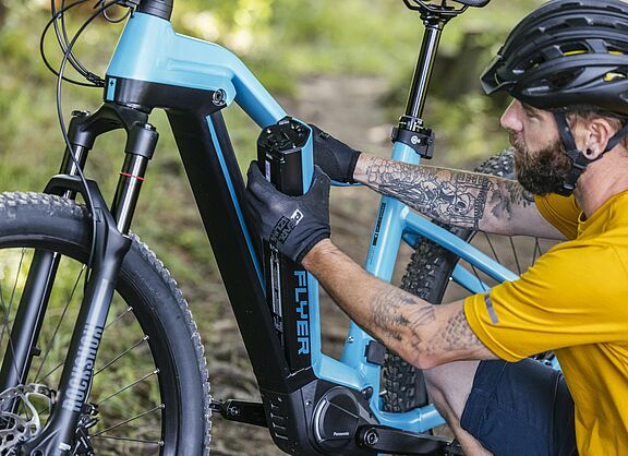 Ein Mann in gelbem Trikot und schwarzem Fahrradhelm entnimmt einen schwarzen Akku aus dem hellblauen Rahmen seines Pedelecs.